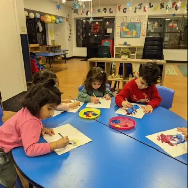 Four young children painting with colorful paints on paper at a round blue table in a classroom.