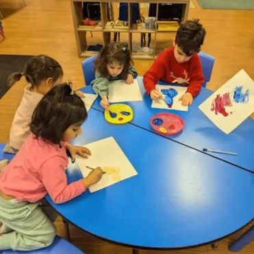 Four young children painting on paper with colorful paints in a vibrant and organized classroom.