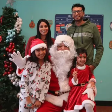 Family posing with Santa Claus in a festive classroom decorated with a Christmas tree, ornaments, and holiday-themed outfits.